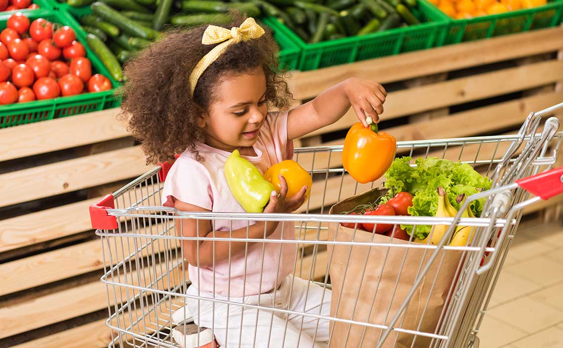 Young child in a shopping cart holding bell peppers in a grocery store.
