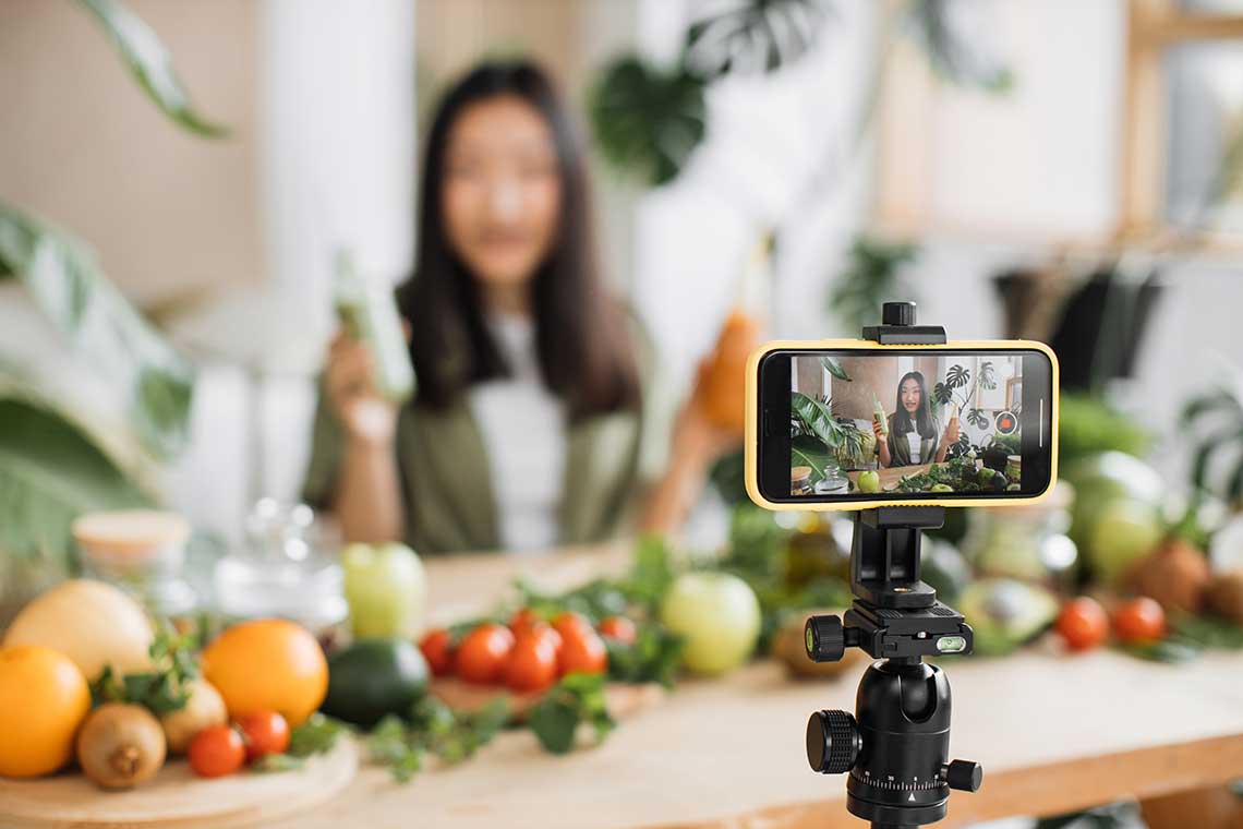 Person recording a video with a smartphone on a tripod, surrounded by fresh vegetables and plants.