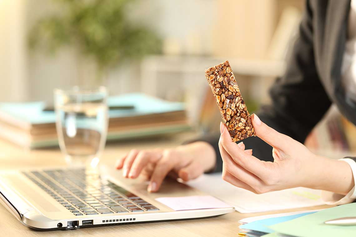 Person holding a granola bar while working on a laptop at a desk.