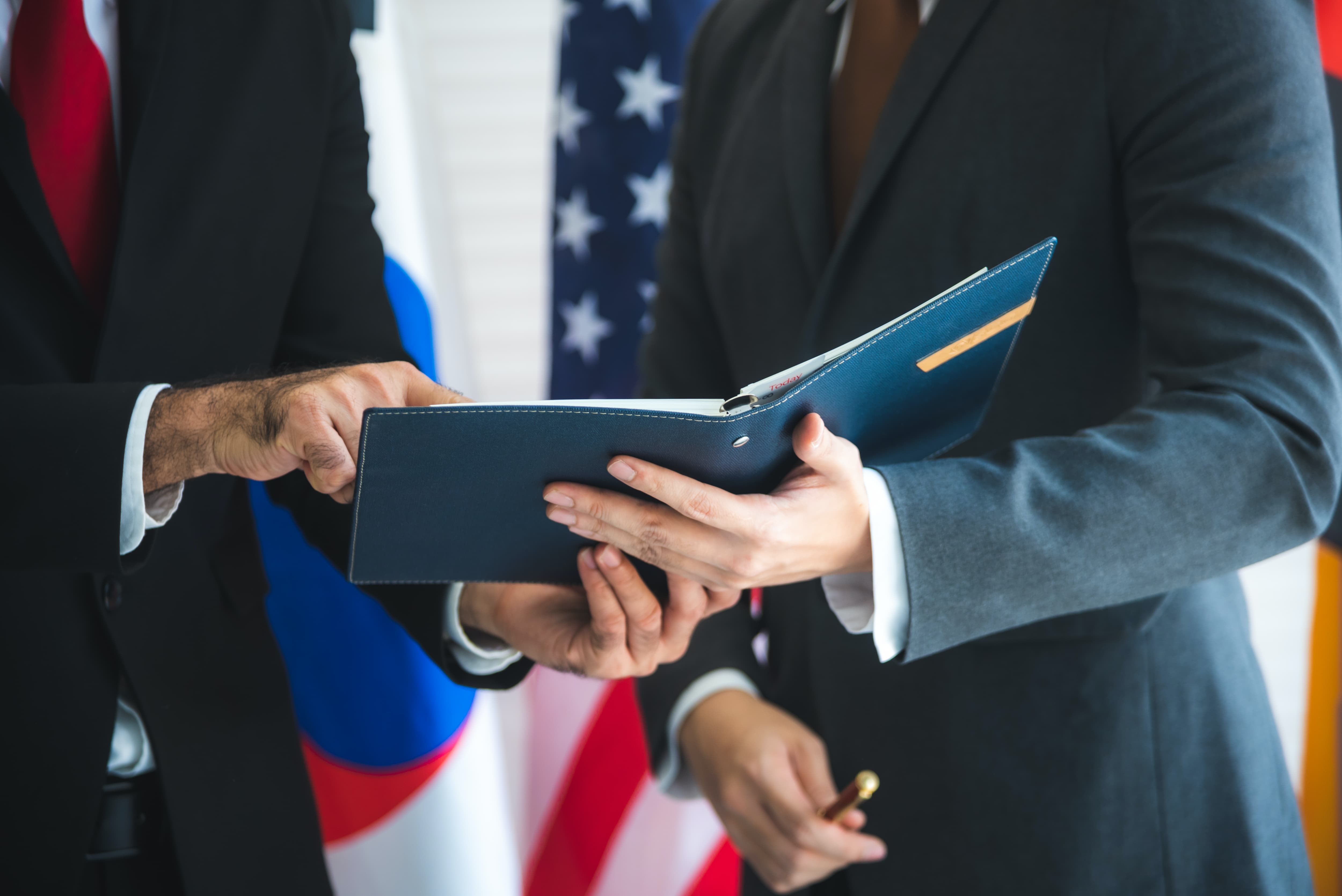Two men in suits look over a proposal while standing in front of flags. Two men in suits look over a proposal while standing in front of flags.