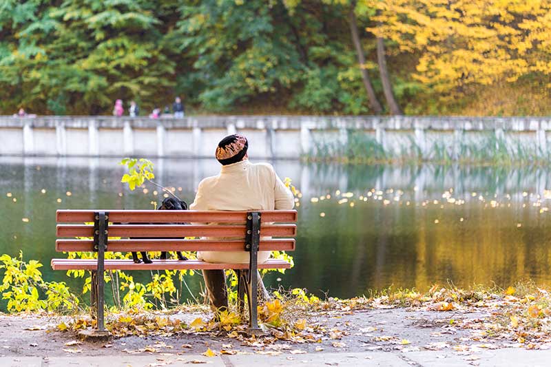 Lonely senior woman with a small black dog sitting on a bench by the autumn lake or river in a city park.