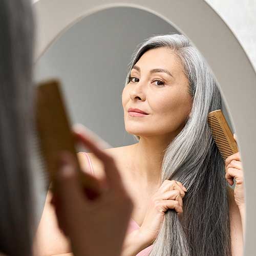 Senior attractive middle 50 years aged Asian woman with gray hair looking at mirror reflection combing tangled gray hair.