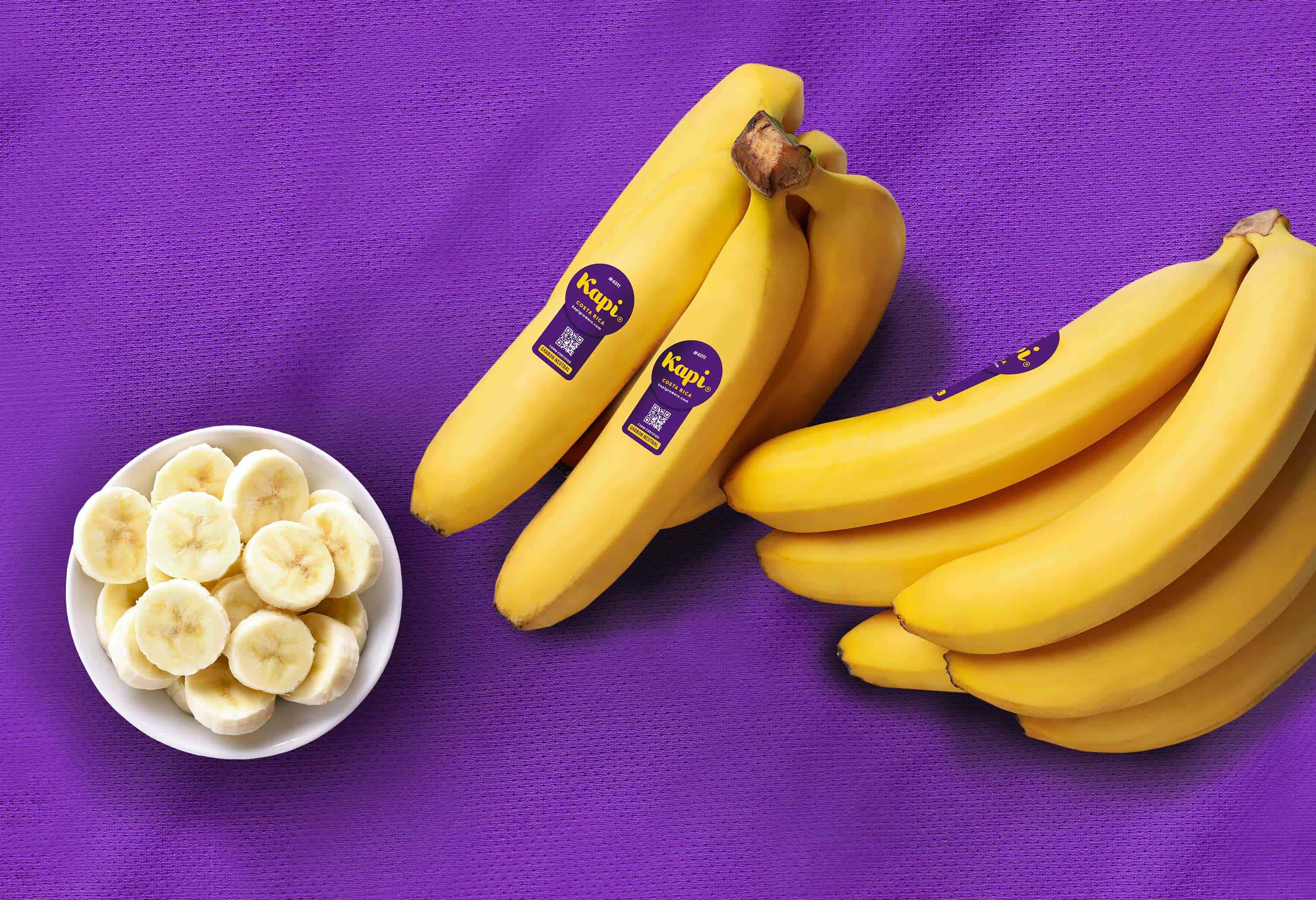 Bright two bunches of Kapi bananas on a bright purple cloth with a bowl of sliced bananas to the left.
